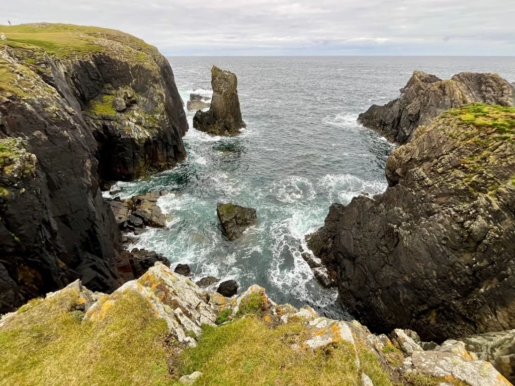 Weitwinkel-Ansicht der zerklüfteten Küstenlinie mit einer markanten Felsnadel (Stack), die isoliert im türkisblauen Wasser vor den hohen Klippen steht.