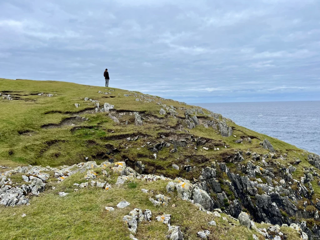 Eine einsame Silhouette eines Menschen steht am Rande einer gewaltigen, flachen Steilklippe über dem Meer unter einem wolkenverhangenen Himmel.