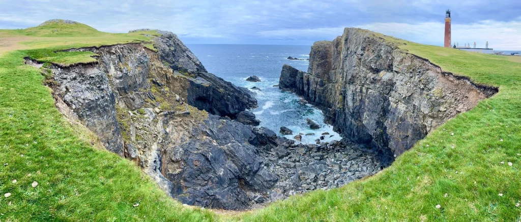 Eine weite Panorama-Aufnahme einer zerklüfteten Bucht. Steile, teils grasbewachsene Klippen fallen zum türkisblauen Atlantik ab, in dem schroffe Felsinseln aus der Brandung ragen.