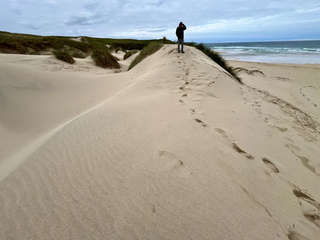 Eine Person steht auf dem Kamm einer hohen, hellen Sanddüne und blickt auf den Ozean hinaus. Im Vordergrund sind Fußspuren im Sand zu sehen, die über den Dünenrücken führen. Im Hintergrund erstreckt sich der Strand und die Brandung unter einem weiten, grauen Wolkenhimmel.