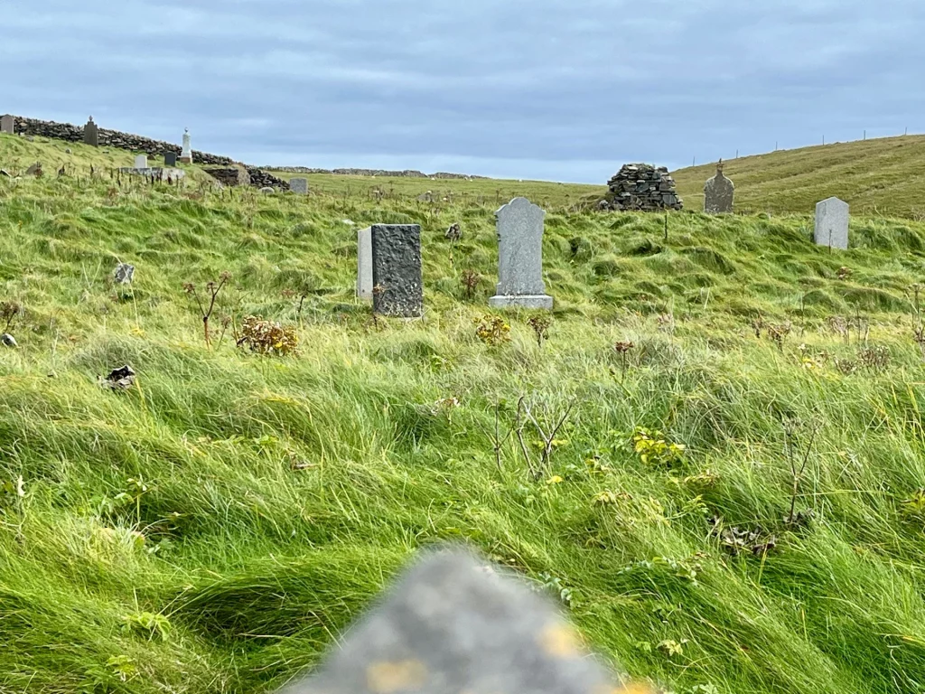 Ein historischer Friedhof in Hanglage mit verstreuten, grauen Grabsteinen, die aus dem hohen, vom Wind gebeugten Gras ragen.