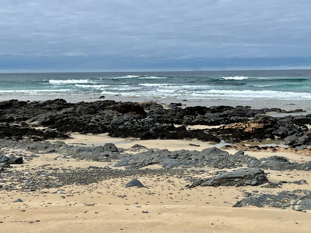 Ein weiter Blick über einen rauen Küstenabschnitt. Im Vordergrund dunkle Felsformationen und Sand, dahinter die heranrollende Brandung des blauen Ozeans unter einem bedeckten Himmel.