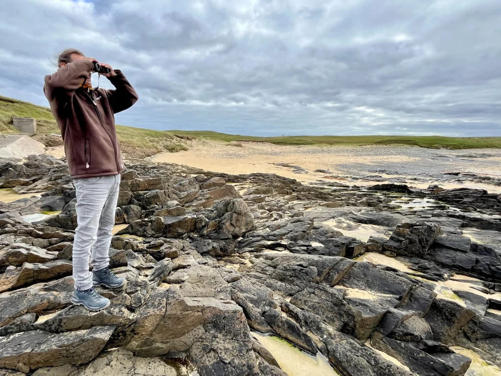 Ein Mann steht in Wanderkleidung auf einer zerklüfteten, dunklen Felsküste und blickt durch ein Fernglas in die Ferne. Im Hintergrund erstreckt sich ein einsamer Sandstrand unter einem weitläufigen, wolkenverhangenen Himmel.