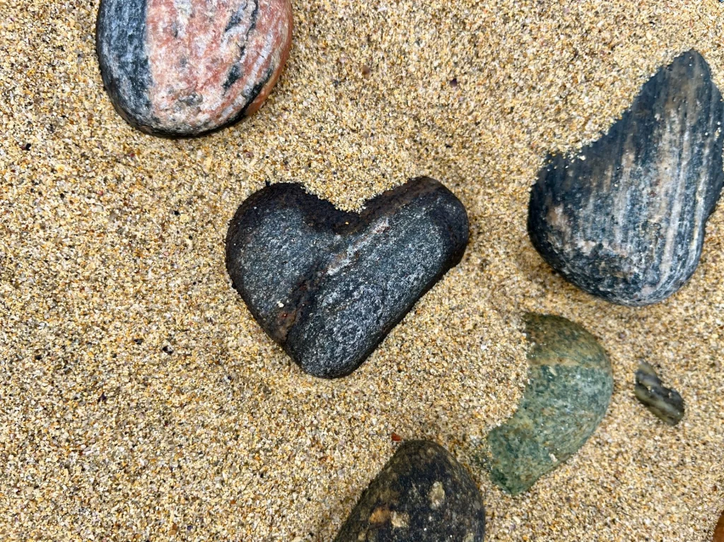 Eine Draufsicht auf groben Sand, in dem mehrere Kieselsteine liegen. Besonders auffällig ist ein dunkler, glatter Stein in der Mitte, der fast perfekt die Form eines Herzens hat. Daneben liegen ein rötlich gemusterter und ein bläulicher Stein.