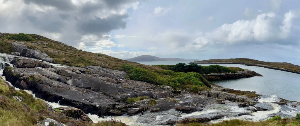 Panorama-Aufnahme einer wilden schottischen Küstenlandschaft. Im Vordergrund fließt ein kleiner Wasserfall über dunkle, bemooste Felsen in ein Loch. Im Hintergrund sieht man sanfte, karge Hügelketten und die Meeresausläufer unter einem dramatisch bewölkten Himmel.