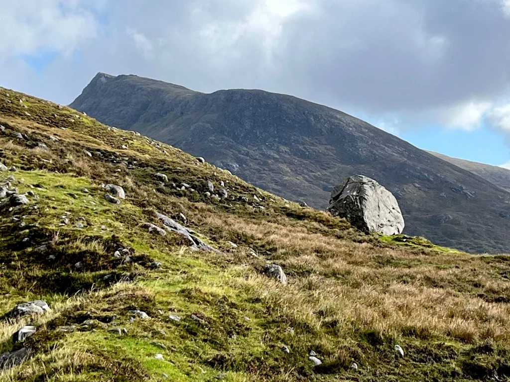 Ein massiver, grauer Findling liegt markant auf einem grasbewachsenen Berghang vor einem steilen Gipfel im Hintergrund.