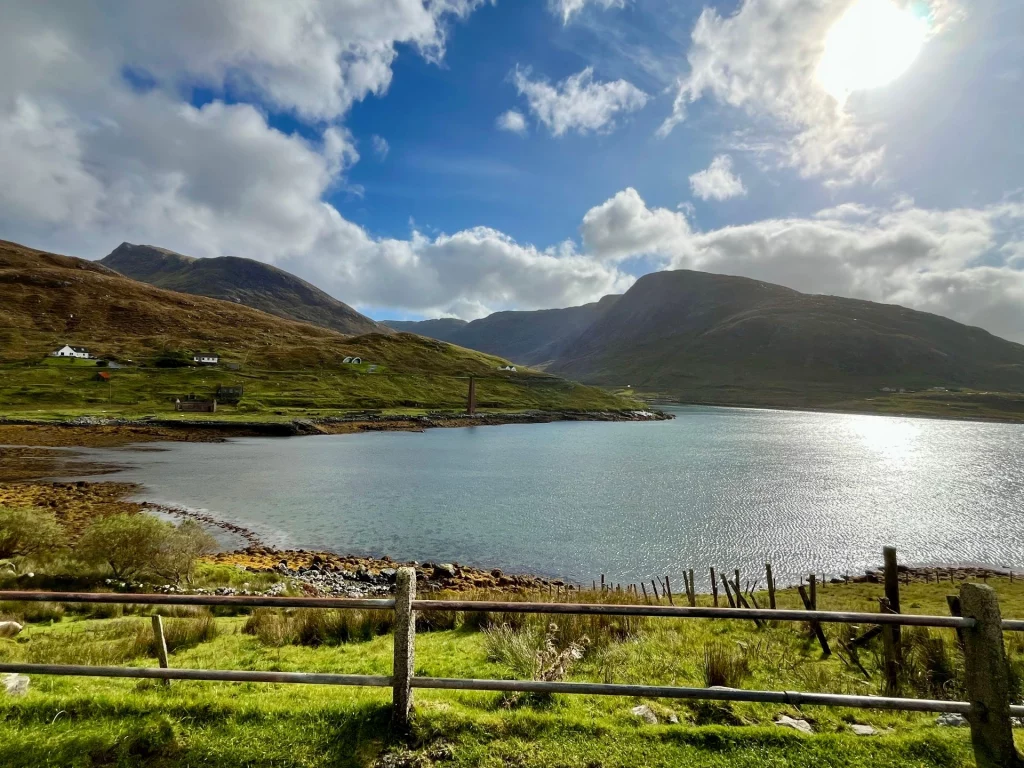 Weite Ansicht einer Meeresbucht, umgeben von dunklen Bergen unter einem dramatischen Wolkenhimmel mit durchscheinender Sonne.