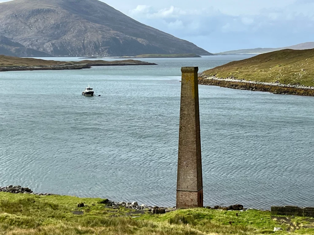 Ein einsamer, hoher Backsteinschornstein (Überrest einer alten Industrieanlage) am Ufer einer blauen Meeresbucht vor bergiger Kulisse.