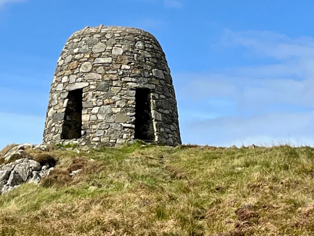 Ein historischer, kuppelförmiger Rundbau aus trocken geschichteten Natursteinen auf einer Anhöhe unter blauem Himmel.