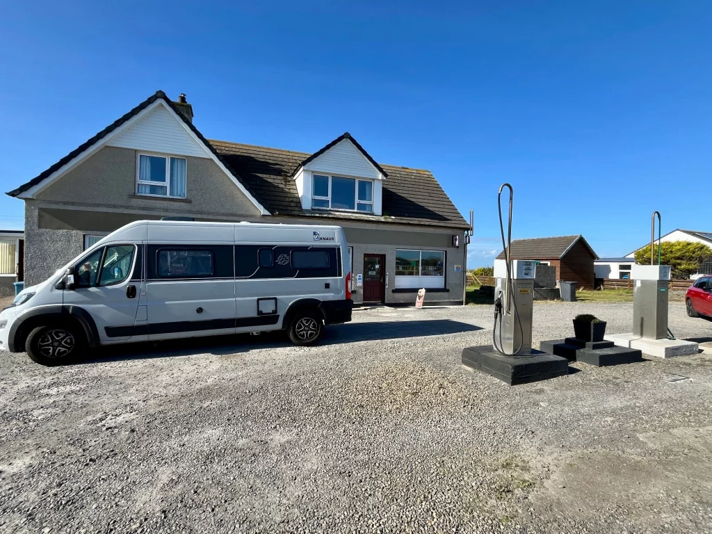 Ein silberner Kastenwagen (Camper) parkt auf einem Schotterplatz vor einer kleinen, ländlichen Tankstelle in John o’ Groats. Im Vordergrund stehen zwei freistehende Zapfsäulen. Dahinter befindet sich ein graues Wohnhaus mit weißem Giebel, in dem ein kleiner Laden mit Poststelle untergebracht ist. Der Himmel ist strahlend blau und wolkenlos.