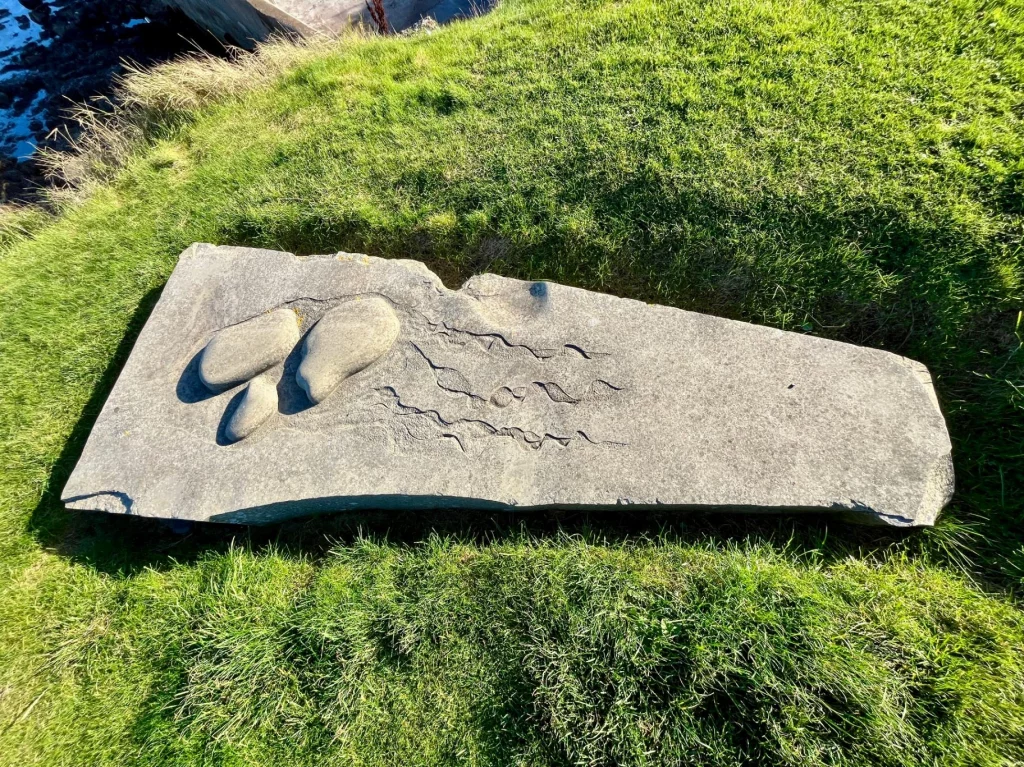 Eine große, flache Steinplatte liegt im grünen Gras. In den Stein sind drei Fußabdrücke unterschiedlicher Größe sowie Wellenlinien eingraviert, die den Weg zum Meer symbolisieren.