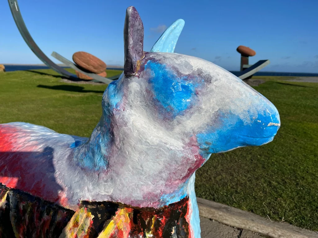 Nahaufnahme einer farbenfroh bemalten Skulptur, die einen Tierkopf (ähnlich einem Schaf oder Kalb) darstellt. Der Kopf ist in Weiß- und Blautönen gehalten, während der Körper rote und dunkle Muster aufweist. Im Hintergrund erstreckt sich eine grüne Wiese mit einer modernen Metall- und Steininstallation unter einem klaren blauen Himmel.