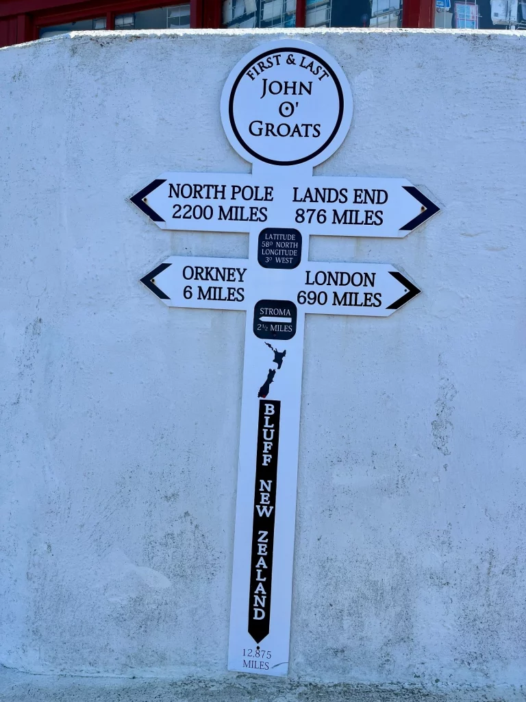Ein weißer Wegweiser in John O'Groats vor einer hellen Wand. Die Schilder zeigen Entfernungen an, darunter zum Nordpol (2200 Meilen) und nach Land's End (876 Meilen).