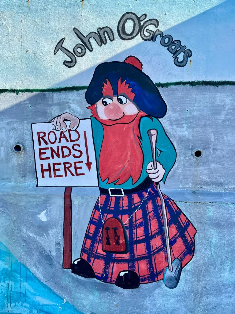 Ein farbenfrohes Wandgemälde eines bärtigen Schotten in traditionellem Kilt und blauer Mütze. Er hält ein Schild mit der Aufschrift „Road ends here“ (Straße endet hier) unter dem Schriftzug „John O'Groats“.