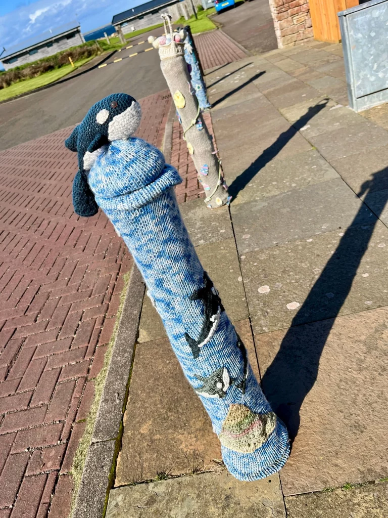 Ein Metallpfosten am Straßenrand, der komplett mit einer hellblauen, gestrickten Hülle („Yarn Bombing“) überzogen ist. Auf der Spitze thront eine kleine, gestrickte Orca-Figur.