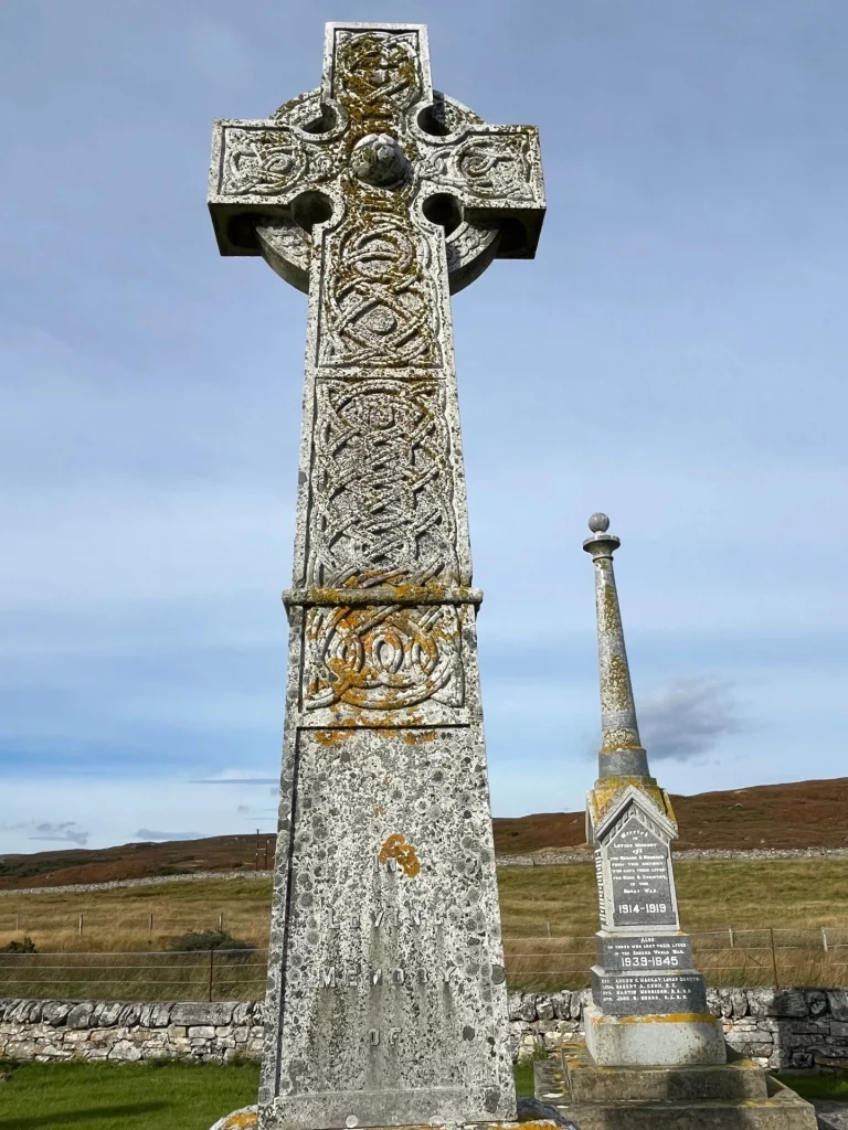 Nahaufnahme eines monumentalen keltischen Hochkreuzes aus grauem Stein auf einem Friedhof in Schottland. Das Kreuz ist über die gesamte Fläche mit traditionellen, komplexen Knotenmustern und Flechtwerk-Reliefs verziert, die teilweise von gelben Flechten bewachsen sind. Im Hintergrund ist ein weiteres, schlankeres Denkmal in Form eines Obelisken zu sehen, das an die Gefallenen der Weltkriege (1914–1919 und 1939–1945) erinnert. Das Ensemble steht vor einer kargen Hügellandschaft unter einem hellen, leicht bewölkten Himmel.