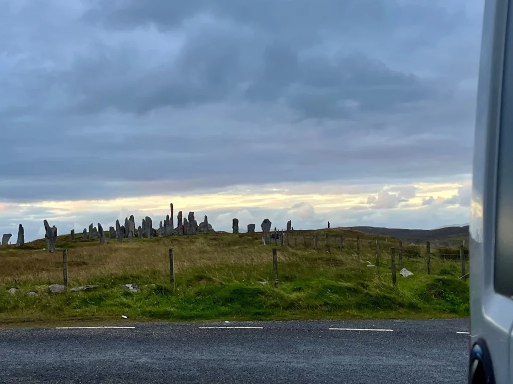 Fernansicht der stehenden Steine von Callanish (Callanish Standing Stones) am Horizont bei dämmrigem Licht. Die Silhouette der Megalithen hebt sich gegen den hellen Streifen am bewölkten Abendhimmel ab.