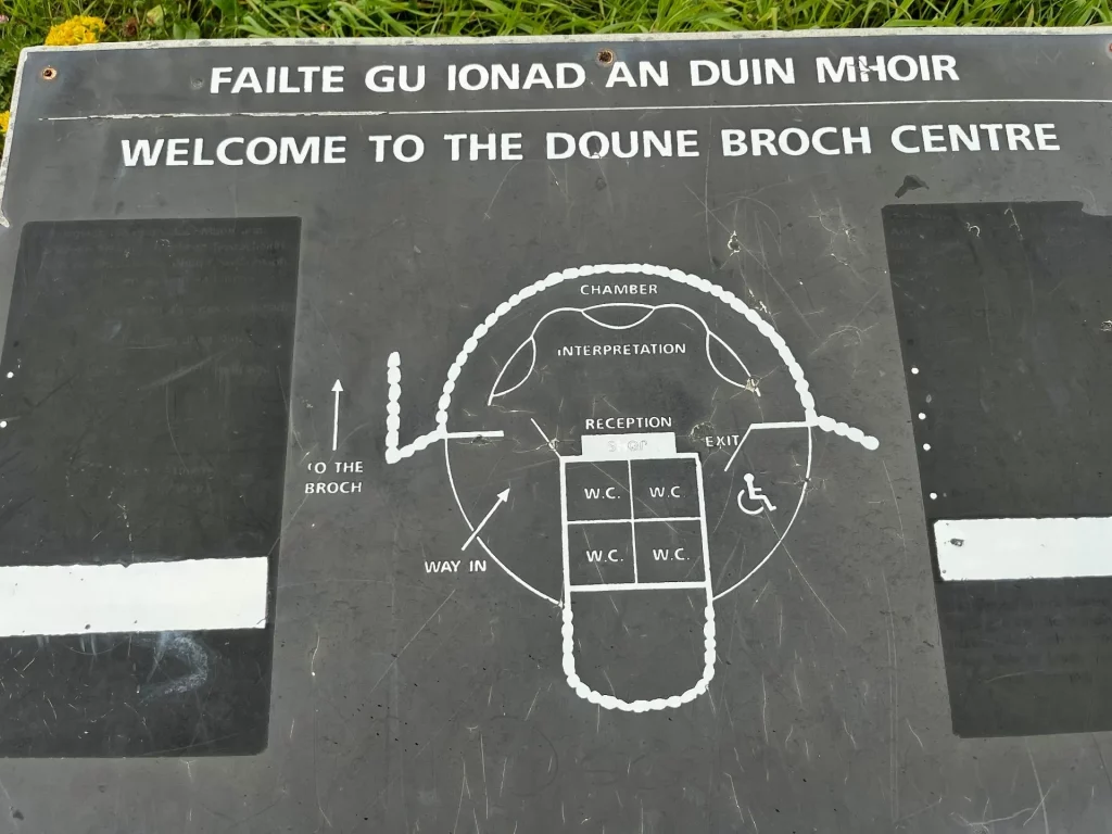 Eine dunkle Informationstafel mit der Aufschrift „Welcome to the Doune Broch“ zeigt einen weißen Grundriss des Geländes und markiert barrierefreie Zugänge.