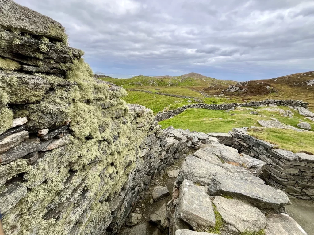 Blick entlang der oberen Mauerkante des Brochs. Die massiven, mit Flechten bewachsenen Trockenmauersteine führen den Blick in die weite, hügelige Landschaft unter einem bewölkten Himmel.