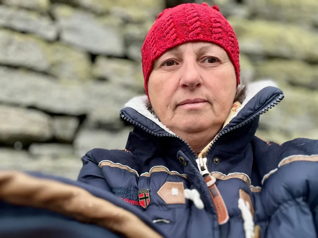 Eine Frau mit roter Strickmütze und blauer Winterjacke blickt vor dem Hintergrund der historischen Trockenmauer von Dun Carloway direkt in die Kamera.