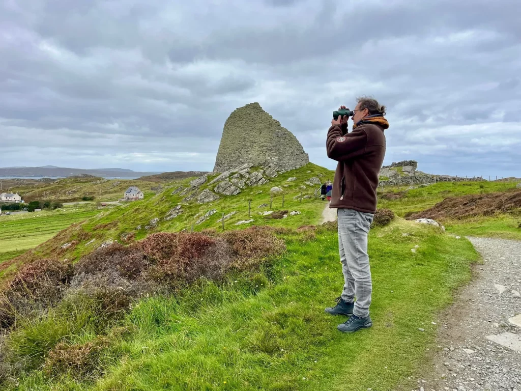 Ein Mann beobachtet mit einem Fernglas den historischen Dun Carloway Broch auf einem grünen Hügel der Isle of Lewis.