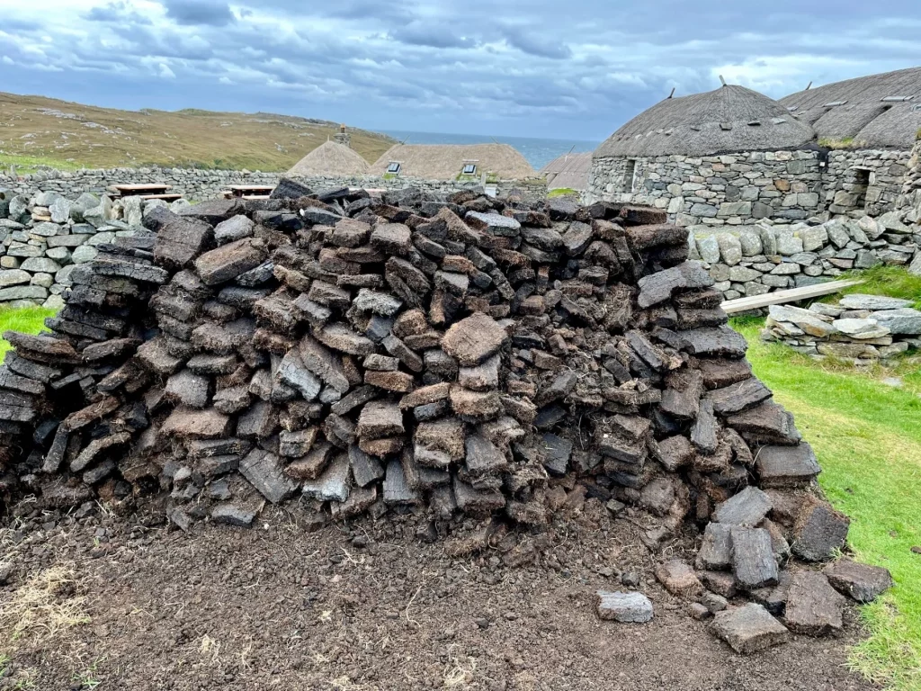 Ein großer Haufen getrockneter Torfsoden im Vordergrund mit den steinernen Blackhouses von Na Gearrannan im Hintergrund unter bewölktem Himmel.