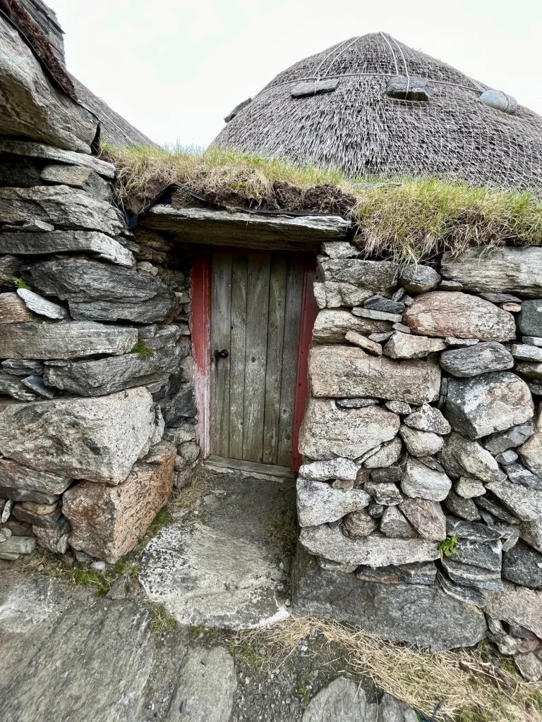 Nahaufnahme einer niedrigen, hölzernen Eingangstür eines Blackhouses in Na Gearrannan, eingefasst in eine massive Trockensteinmauer mit einem grasbewachsenen Dachvorsprung.