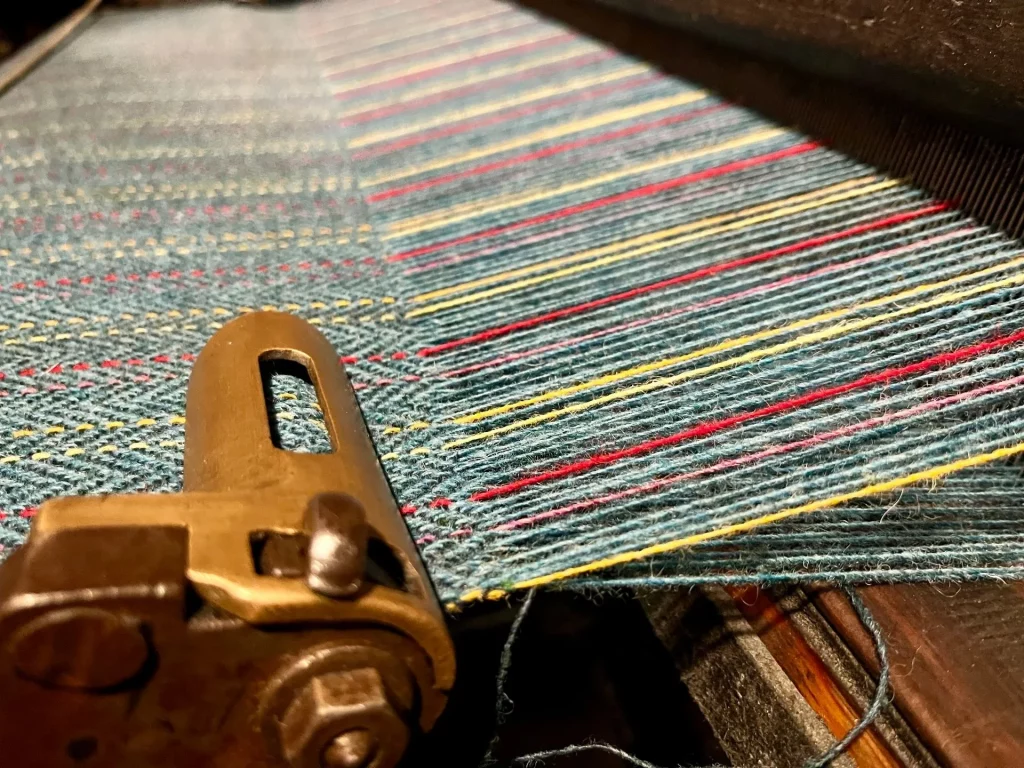 Nahaufnahme eines mechanischen Webstuhls während der Arbeit, auf dem ein bunt gestreifter Harris Tweed Stoff mit gelben, roten und blauen Fäden entsteht.