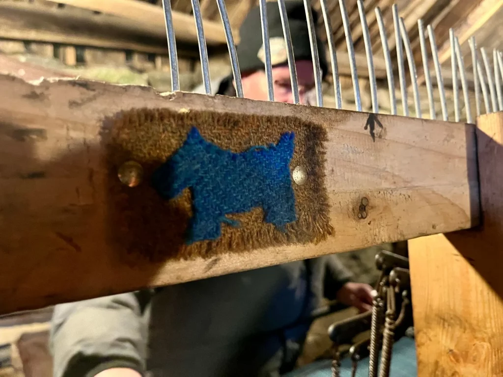 Detailaufnahme eines hölzernen Wollkamms mit langen Metallzinken, auf dem ein kleines blaues Stoffstück in Form eines Hundes (Terrier) befestigt ist.