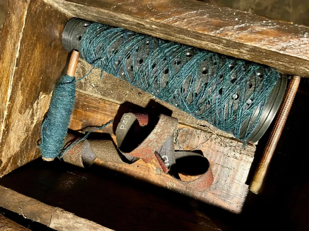 Eine traditionelle Holzspule, die stramm mit blaugrünem Garn umwickelt ist, liegend in einer rustikalen Holzvorrichtung.