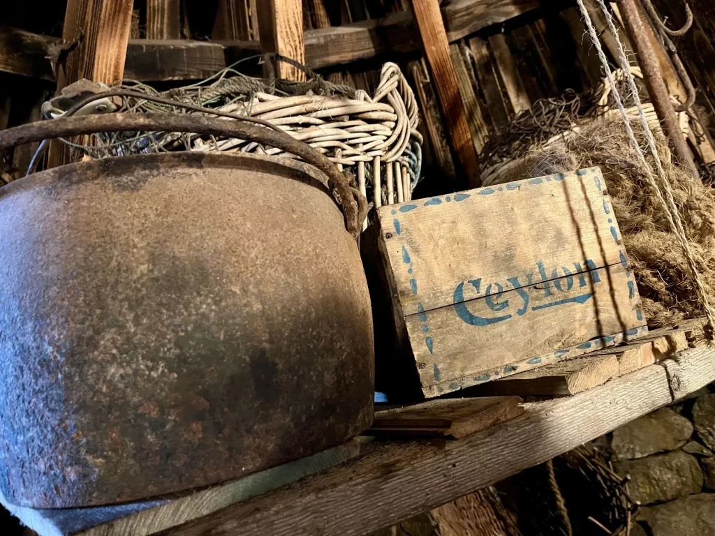 Stillleben in einem Blackhouse mit einem alten gusseisernen Kochtopf, einem geflochtenen Korb und einer historischen Holzkiste mit der Aufschrift „Ceylon“.