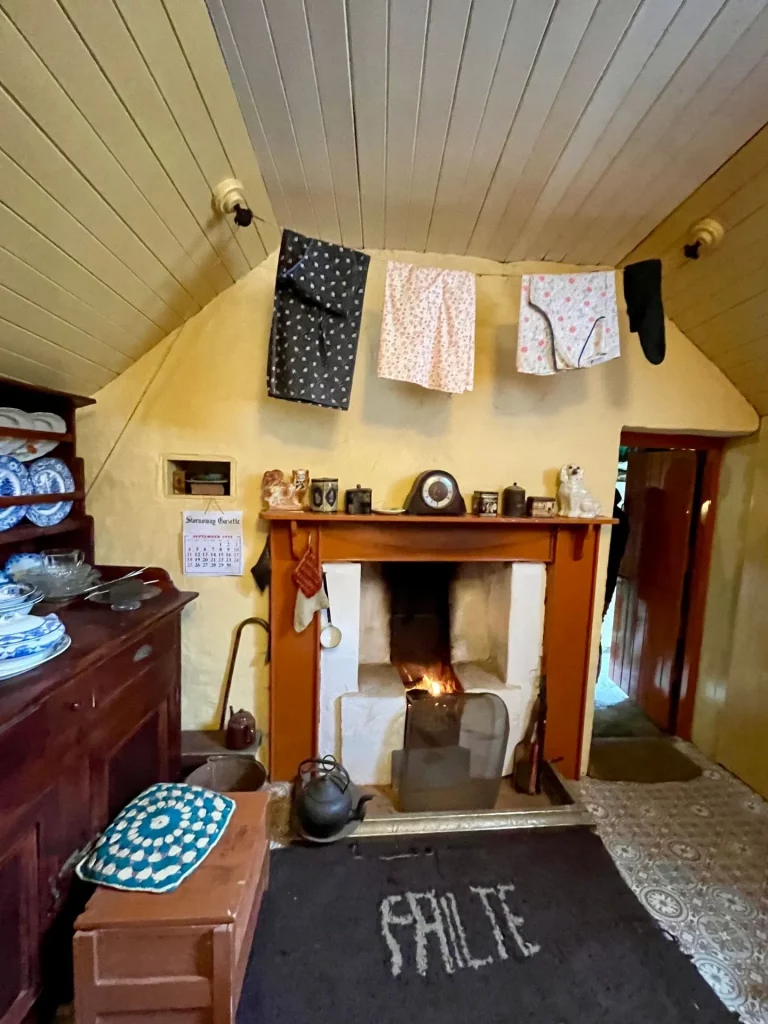 Gemütliches Zimmer mit brennendem Kamin, darüber aufgehängter Wäsche an einer Leine und einer „Failte“-Fußmatte (Gälisch für Willkommen) vor dem Feuer.