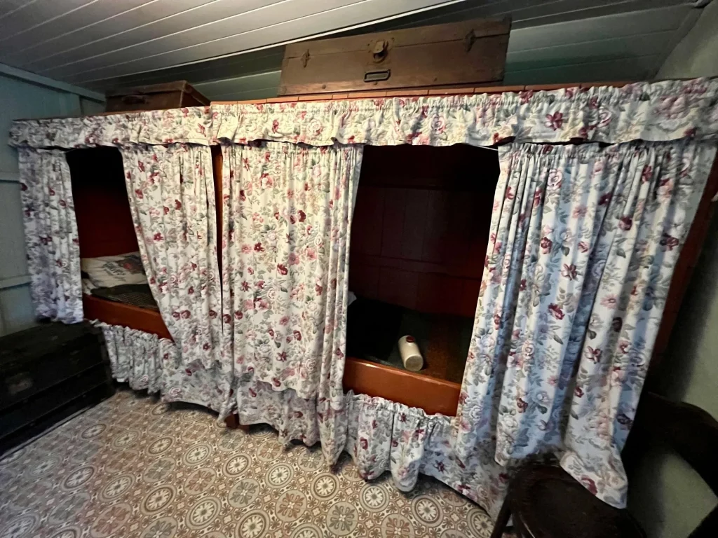 Ein traditionelles schottisches Alkovenbett (Box Bed), das hinter blumig gemusterten Vorhängen in die Wand eingebaut ist, mit einem hölzernen Koffer auf dem oberen Sims.