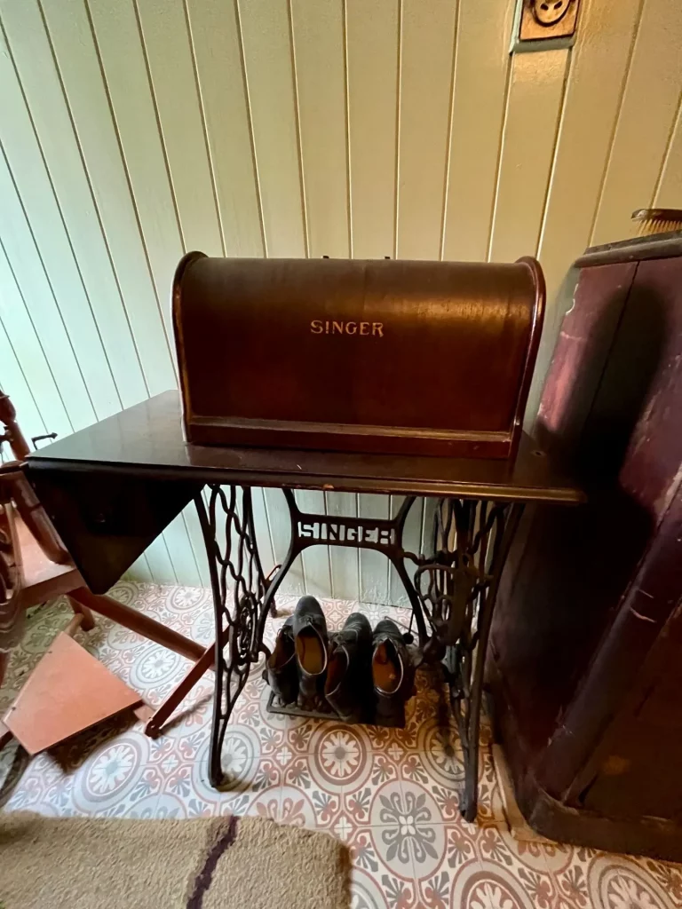 Eine antike Singer-Tretnähmaschine aus dunklem Holz und schwarzem Gusseisen, die vor einer hellen Holzwand auf einem gemusterten Linoleumfußboden steht.
