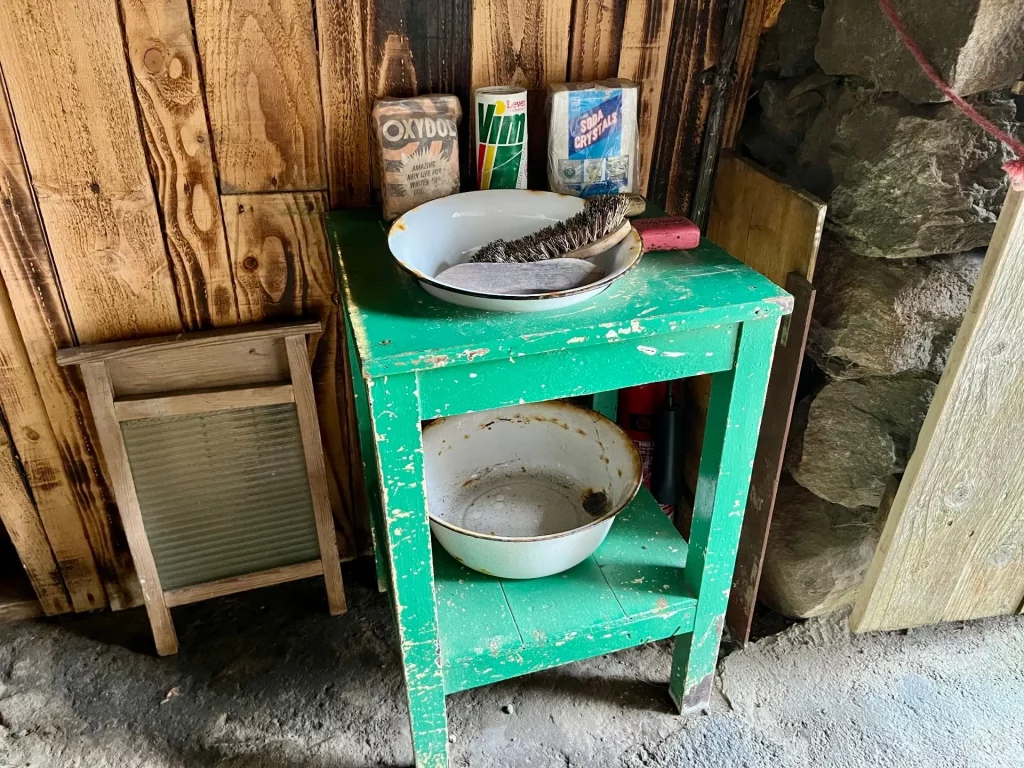 Ein einfacher, grün gestrichener Holztisch mit einer weißen Emaille-Waschschüssel und einer Seifenschale in einer spärlich beleuchteten Kammer.