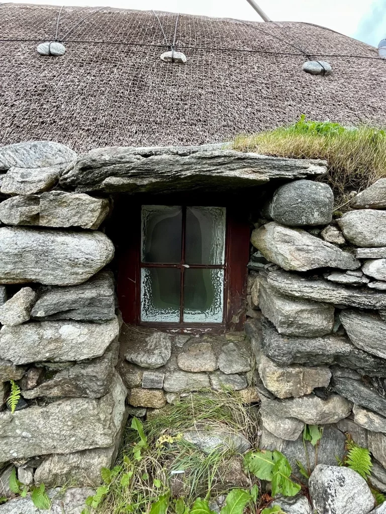 Ein kleines, tief in die dicke Natursteinmauer eingelassenes Sprossenfenster eines Blackhouses mit einem schweren Steinsturz und darüberliegendem Reetdach, das mit Steinen beschwert ist.