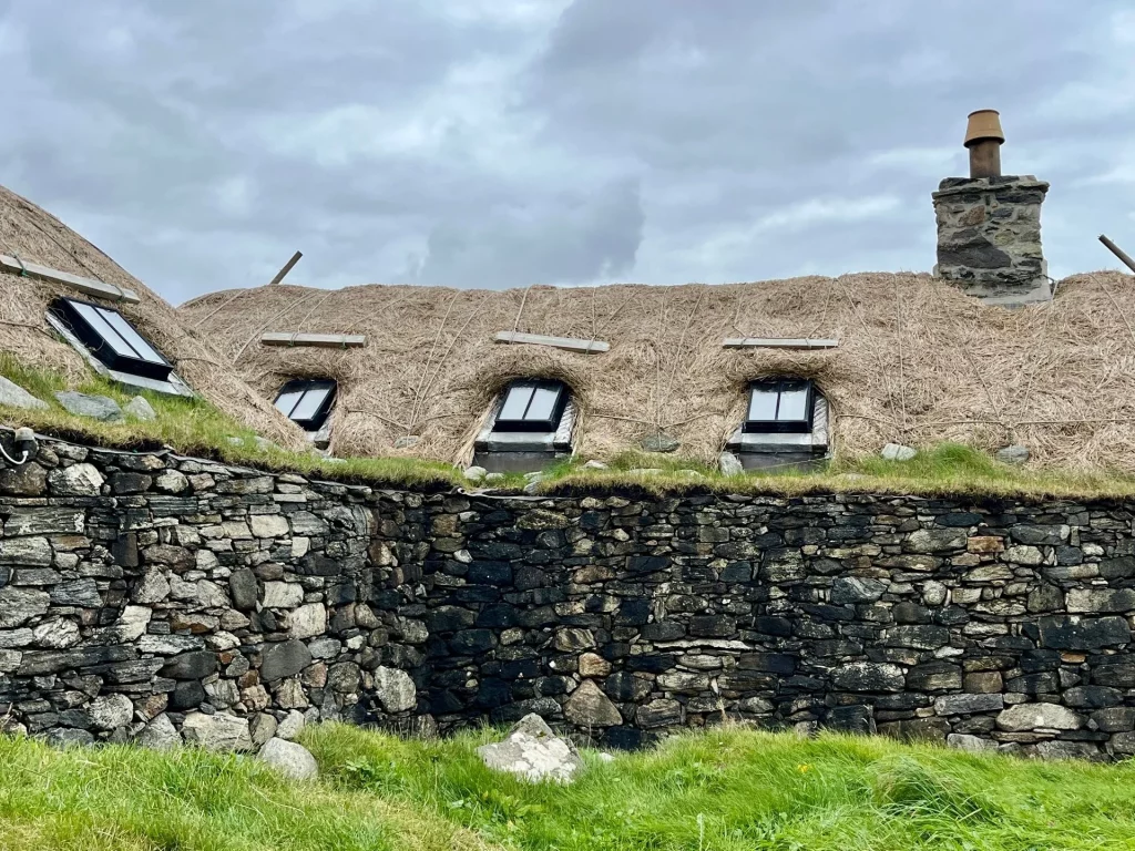 Nahaufnahme eines reetgedeckten Blackhouse-Daches mit modernen Dachfenstern, die in das traditionelle Stroh eingelassen sind, über einer rustikalen Trockensteinmauer.