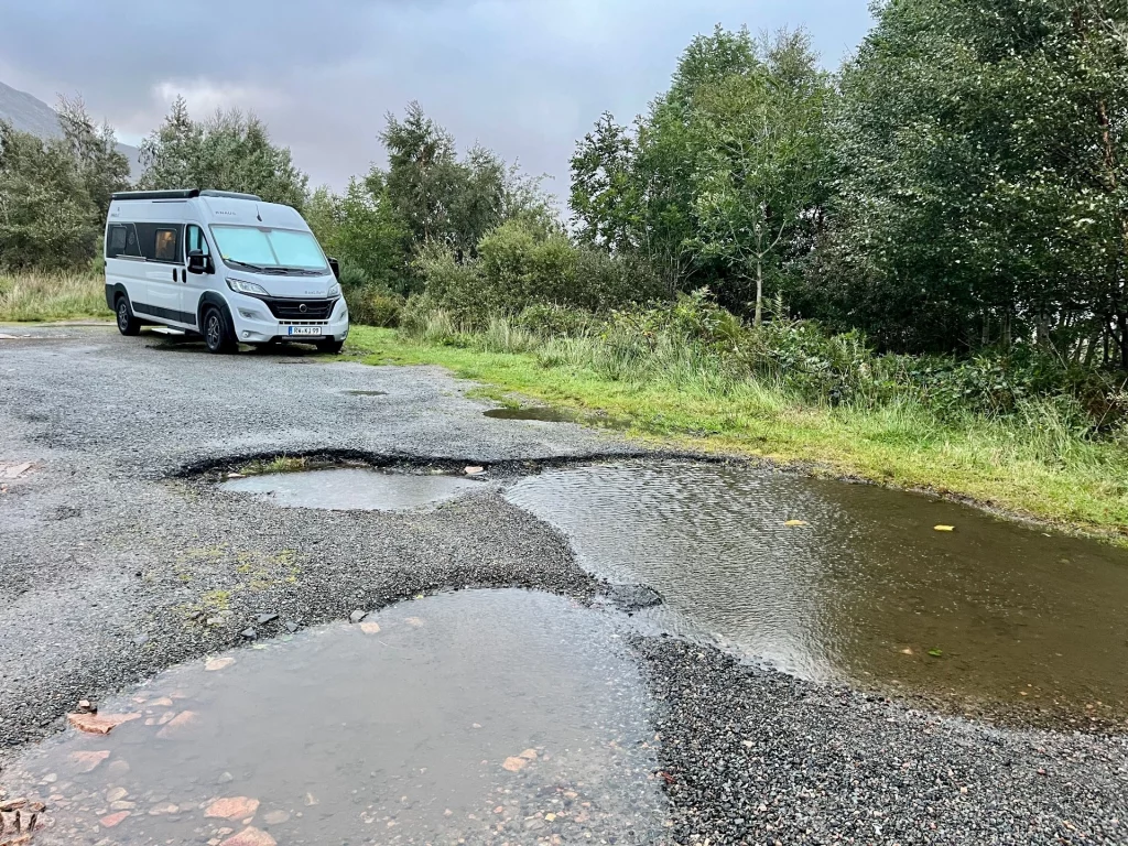 Ein hellgrauer Campervan steht auf einem geteerten Platz. Im Vordergrund sind mehrere große, wassergefüllte Schlaglöcher zu sehen, die den regnerischen Tag widerspiegeln.