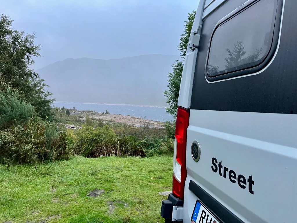 Nahaufnahme der Rückseite eines weißen Kastenwagens mit der Aufschrift „Street“. Im Hintergrund erstreckt sich eine grüne Wiese vor einem nebligen See und Bergen.