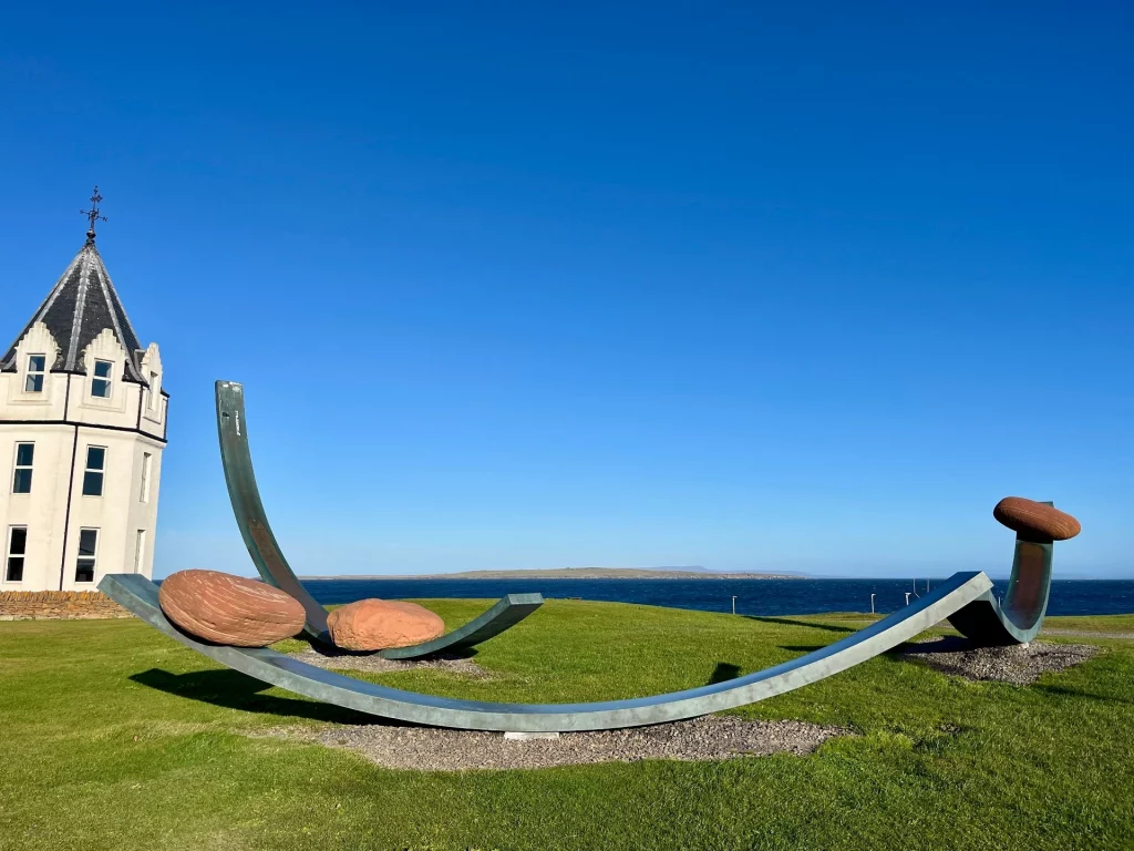 Die Skulptur "Nomadic Boulders" von Julian Meredith: Bronzebögen halten massive rote Sandsteinfelsen auf einer grünen Wiese am Meer.