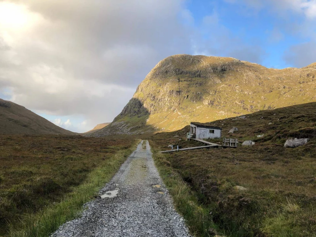 Eine schmale Schotterstraße führt durch eine karge, moosbewachsene Hügellandschaft direkt auf einen markanten, runden Berg zu. Rechts am Wegrand steht eine kleine, einfache weiße Hütte.