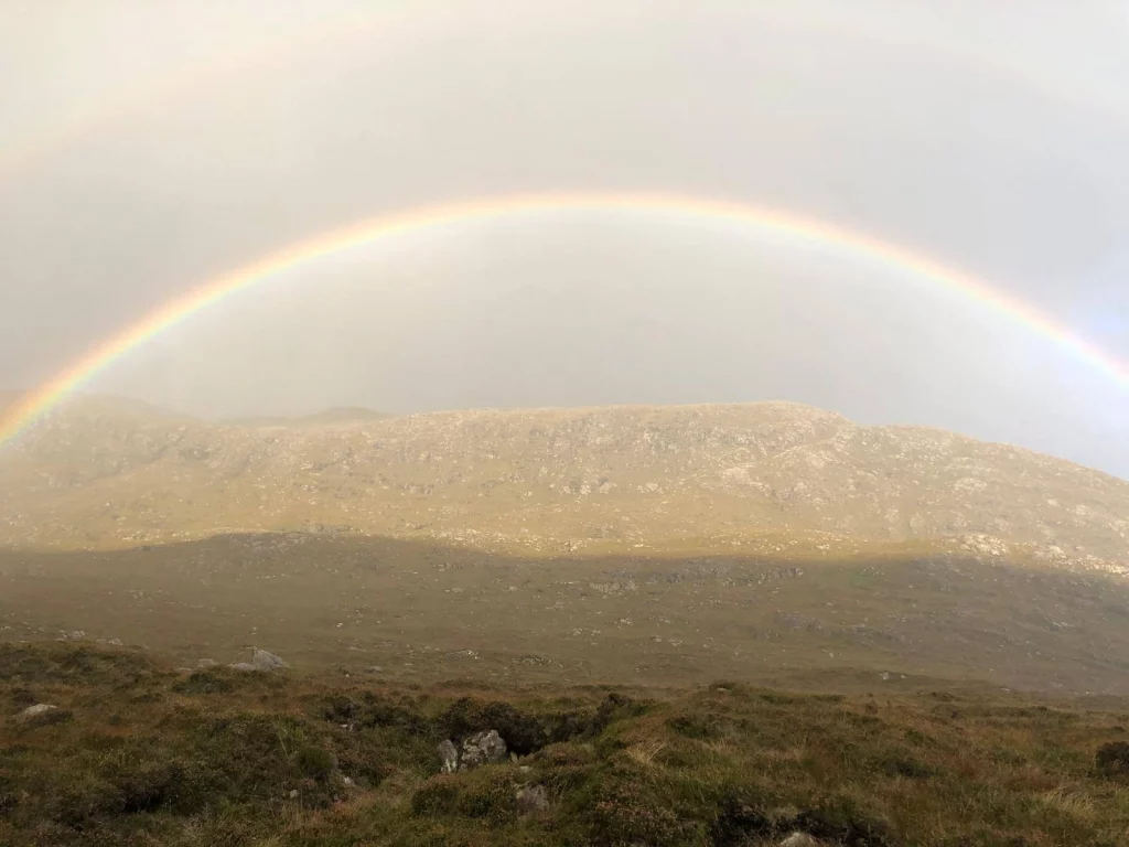 Ein weiter Blick über eine flache, braun-grüne Heide- und Moorlandschaft. Über den entfernten Hügeln spannt sich ein sanfter, vollständiger Regenbogen unter einem wolkenverhangenen Himmel.