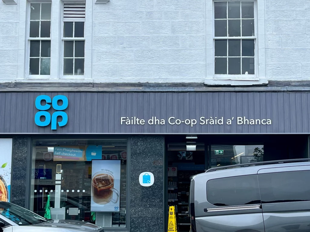 Nahaufnahme der Fassade eines „Co-op“-Supermarkts in Portree mit gälischer Beschriftung „Fàilte dha Co-op Sràid a' Bhanca“ (Willkommen im Co-op der Bank Street) über dem Eingang.