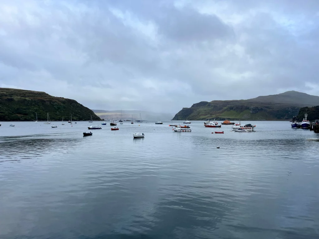 Weite Aufnahme der Bucht von Portree unter einem wolkenverhangenen Himmel; kleine Boote ankern ruhig auf dem spiegelglatten Wasser.
