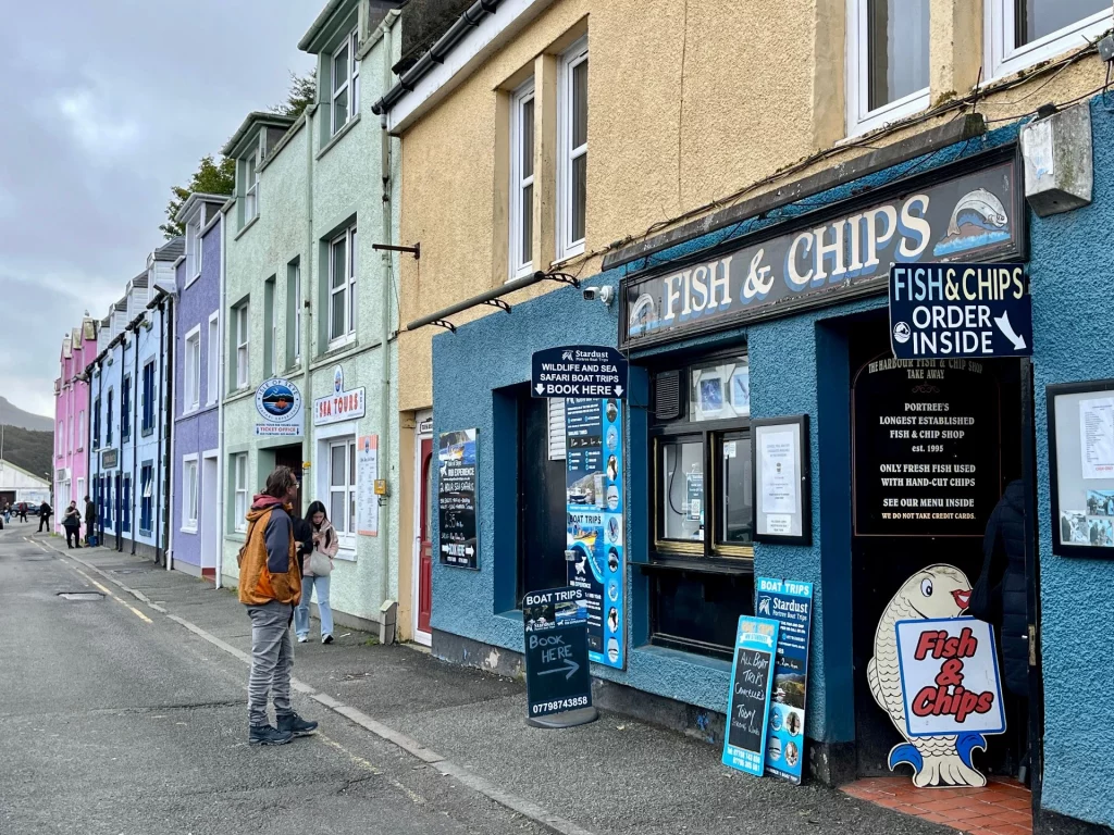 Straßenszene in Portree mit einer Reihe farbenfroher Häuser (rosa, blau, gelb). Im Vordergrund steht ein blauer Imbiss mit der Aufschrift „Fish & Chips“, davor Werbeschilder für Bootstouren und ein Mann in Wanderkleidung.