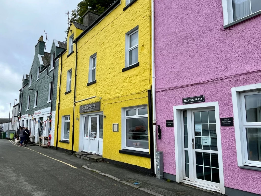 Die berühmte Häuserzeile am Hafen von Portree mit leuchtend gelben und rosa verputzten Fassaden vor einer grauen Straße.