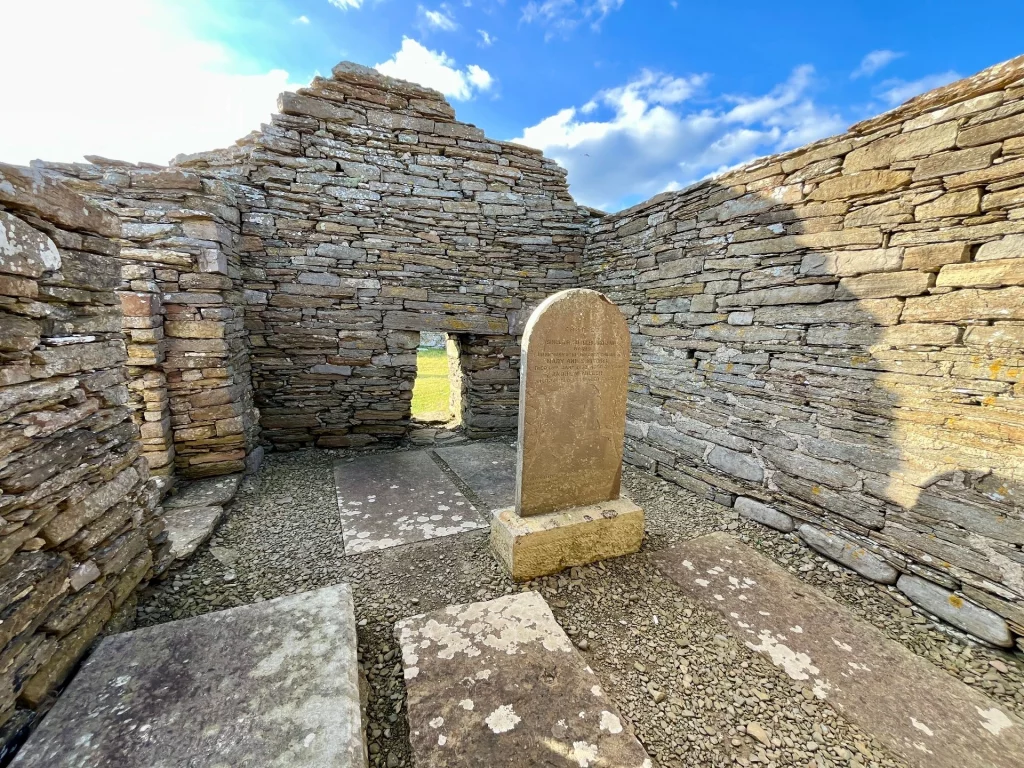 Innenansicht der dachlosen Ruine von St Mary’s Chapel. Man sieht die grob gemauerten Steinwände und mehrere im Boden eingelassene Grabplatten sowie einen aufrecht stehenden Gedenkstein auf einem Kiesboden.