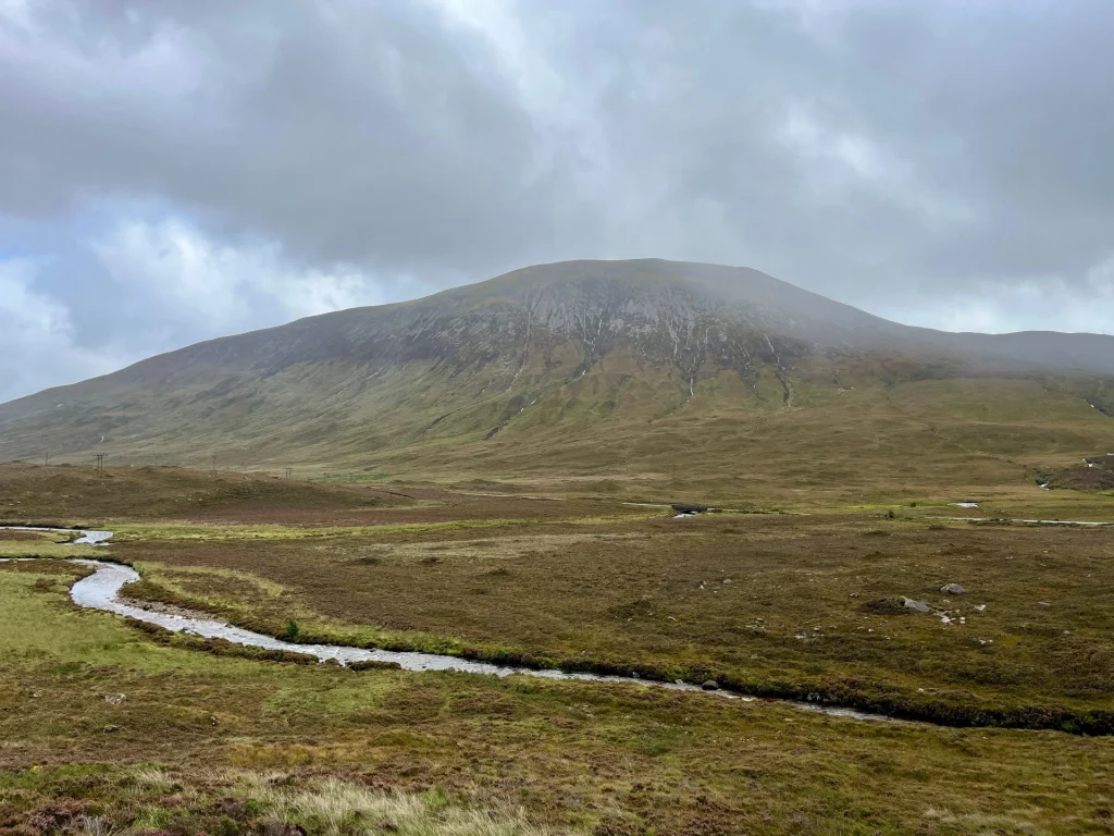 Ein sanfter, grasbewachsener Berg ragt in den grauen, nebligen Himmel. Im Vordergrund schlängelt sich ein schmaler Bach durch die karge, bräunlich-grüne Vegetation der weiten Hügellandschaft.