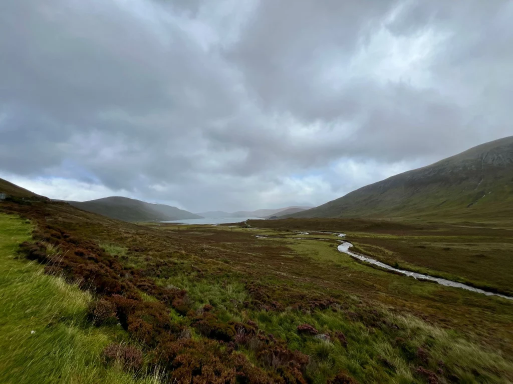 Eine weite, hügelige Moorlandschaft mit rötlich-braunem Heidekraut und grünen Gräsern unter einem dichten, grauen Wolkenhimmel. Ein schmaler, heller Bach schlängelt sich durch das Tal in Richtung einer fernen Bucht, flankiert von sanft ansteigenden Berghängen.