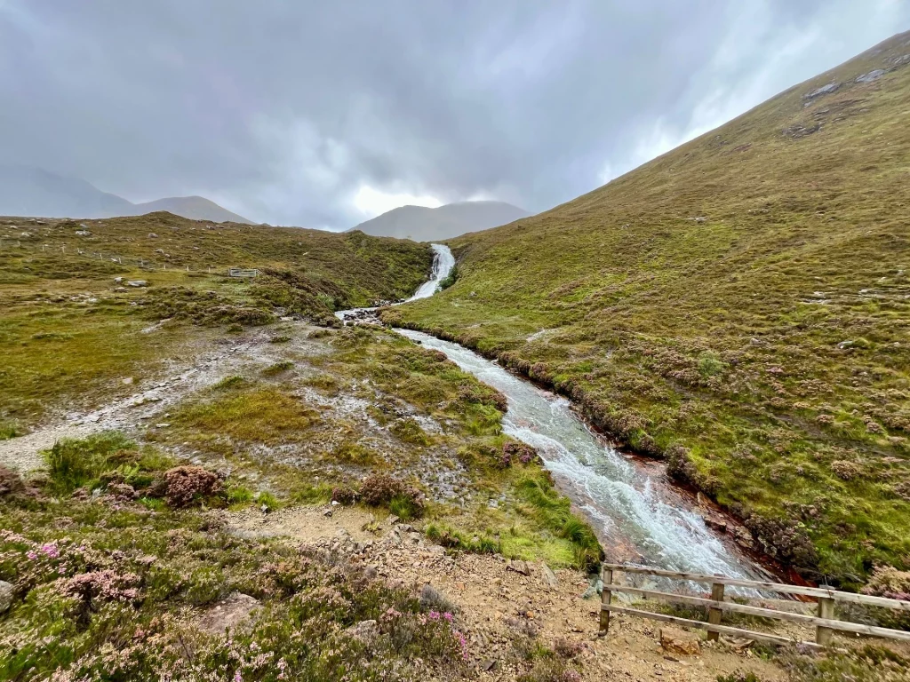 Ein schäumender Bach fließt einen steilen, grünen Hang hinunter und mündet in einen kleinen Wasserfall. Der Blick führt das Tal hinauf in Richtung nebliger Berggipfel unter einer dichten Wolkendecke.